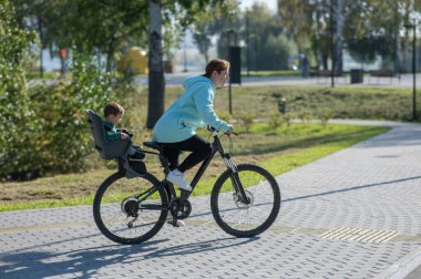 Küçük oğluyla bisiklete binen beyaz bir kadın arkasında çocuk koltuğunda oturuyor.