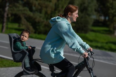 Küçük oğluyla bisiklete binen beyaz bir kadın arkasında çocuk koltuğunda oturuyor.