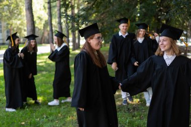 Mezuniyet elbisesi giymiş bir grup mutlu öğrenci dışarıda. Ön planda, iki genç kız birbirini tebrik ediyor. 