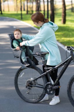 Beyaz bir kadın parkta bir yaşındaki oğluyla bisiklete biniyor. Dikey fotoğraf