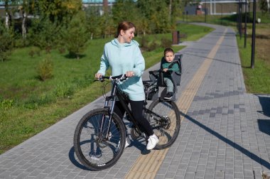 Küçük oğluyla bisiklete binen beyaz bir kadın arkasında çocuk koltuğunda oturuyor.