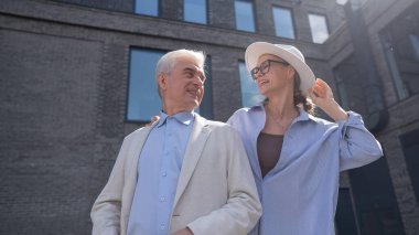 Şapkalı ve gözlüklü bir kadın beyaz ceketli yaşlı bir adama arkadan sarılıyor. Olgun insanların romantik ilişkileri