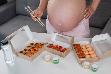 A pregnant woman holds chopsticks and stands at the table with rolls. Close-up of the belly clipart