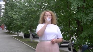 Fazla kilolu beyaz kadın yürürken su içer.