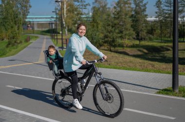Küçük oğluyla bisiklete binen beyaz bir kadın arkasında çocuk koltuğunda oturuyor.