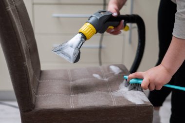Kadın, elektrikli süpürge ve fırça kullanarak kumaş sandalyesini temizliyor.