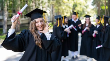 Mezuniyet elbisesi giymiş bir grup mutlu öğrenci dışarıda. Ön planda elinde diploması olan genç bir kız.
