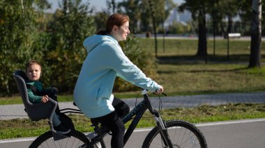Küçük oğluyla bisiklete binen beyaz bir kadın arkasında çocuk koltuğunda oturuyor.