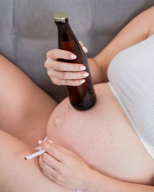 Suratsız hamile kadın kanepede otururken bira içiyor.