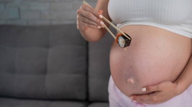 A pregnant woman holds a roll with chopsticks. Close-up of the belly clipart