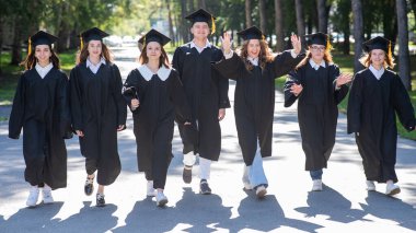 Mezuniyet elbisesi giymiş mutlu gençler. Öğrenciler parkta yürüyor.