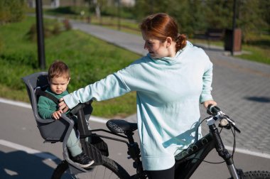 Küçük oğluyla bisiklete binen beyaz bir kadın arkasında çocuk koltuğunda oturuyor.