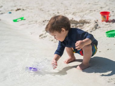 Bir yaşında beyaz bir çocuk deniz kıyısındaki kumda oynuyor.