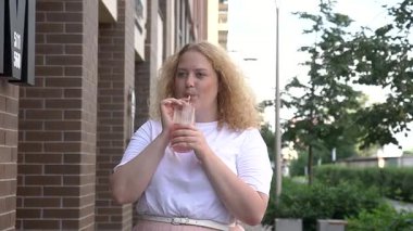 Tombul beyaz bir kadın yürüyor ve pipetten kokteyl içiyor.