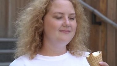 Tombul beyaz bir kadın yürürken dondurma külahı yiyor.