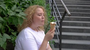 Tombul beyaz bir kadın yürürken dondurma külahı yiyor.