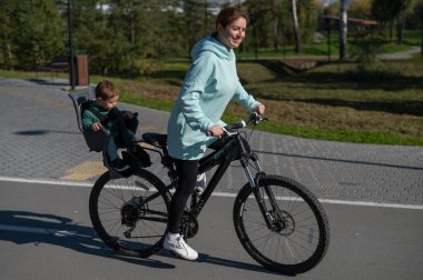 Küçük oğluyla bisiklete binen beyaz bir kadın arkasında çocuk koltuğunda oturuyor.
