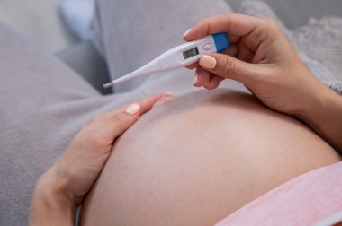 Hamile kadın koltukta otururken sıcaklık ölçer. Karnını kapat.