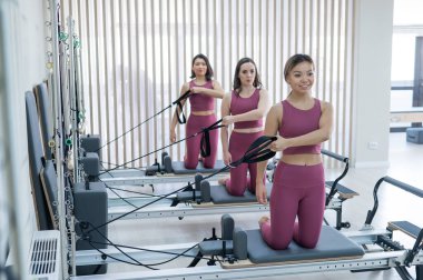 Pembe spor kıyafetli üç kadın ıslah yatağıyla pilates egzersizi yapıyor.