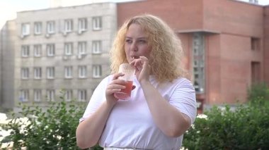 Tombul beyaz bir kadın yürüyor ve pipetten kokteyl içiyor.