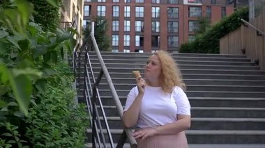 Tombul beyaz bir kadın yürürken dondurma külahı yiyor.