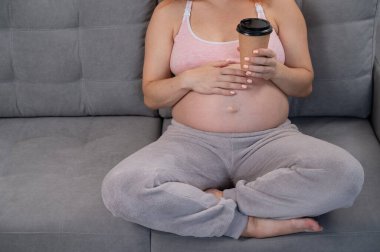 Üç aylık dönemde karnında kahve bardağı tutan yüzü olmayan hamile bir kadın.