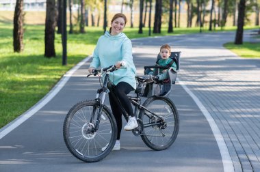 Küçük oğluyla bisiklete binen beyaz bir kadın arkasında çocuk koltuğunda oturuyor.