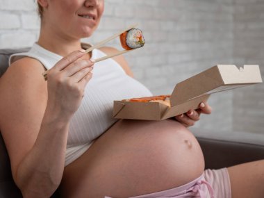 Hamile bir kadın kanepede oturur ve bir kutudan ekmek yer. Yemek teslimatı