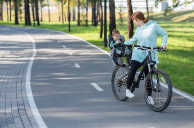 Küçük oğluyla bisiklete binen beyaz bir kadın arkasında çocuk koltuğunda oturuyor.