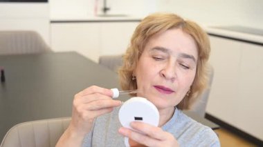 Yaşlı beyaz bir kadın yüzüne serum uyguluyor.