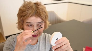 Yaşlı beyaz bir kadın kirpiklerine rimel sürüyor ve küçük bir aynaya bakıyor.