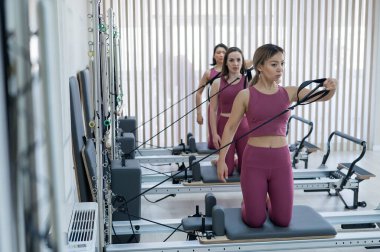 Pembe spor kıyafetli üç kadın ıslah yatağıyla pilates egzersizi yapıyor.