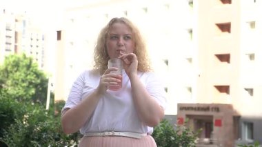 Tombul beyaz bir kadın yürüyor ve pipetten kokteyl içiyor.