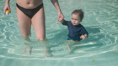 Bir yaşında beyaz bir çocuk annesiyle yüzme havuzunda el ele yürür.