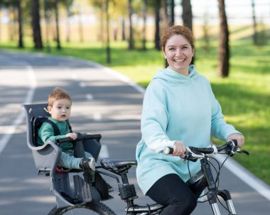 Küçük oğluyla bisiklete binen beyaz bir kadın arkasında çocuk koltuğunda oturuyor.
