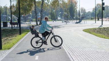 Beyaz kadın bisiklete biniyor. Yanında da oğlu var. Parkta çocuk koltuğunda oturuyor.