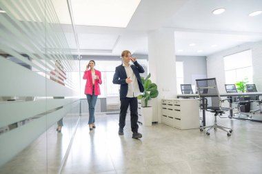 İki meslektaş ofiste yürüyor ve telefonda konuşuyorlar.