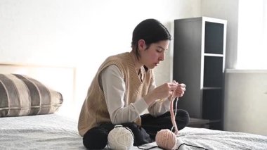 Genç beyaz kadın yatakta otururken örgü örüyor.