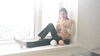 Genç beyaz kadın pencere eşiğinde otururken örgü örüyor.