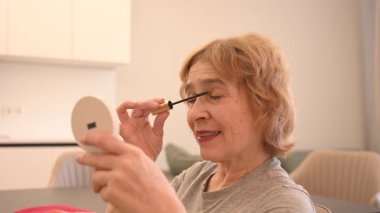 Yaşlı beyaz bir kadın kirpiklerine rimel sürüyor ve küçük bir aynaya bakıyor.