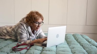Yaşlı, beyaz, pijamalı bir kadın dizüstü bilgisayar kullanarak yatağa uzanıyor.