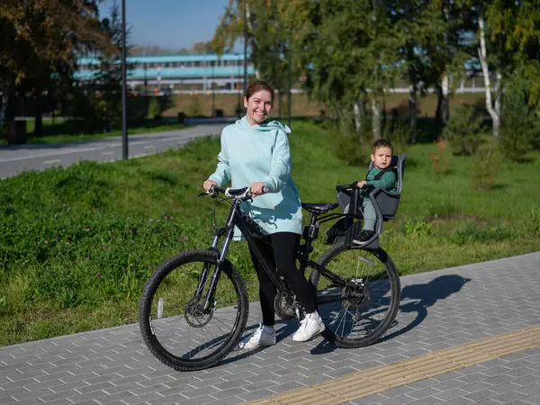 Küçük oğluyla bisiklete binen beyaz bir kadın arkasında çocuk koltuğunda oturuyor.