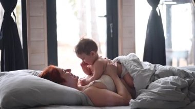 Kızıl saçlı beyaz kadın, panoramik pencereli bir kır evinde bebeğiyle yatakta yatıyor.