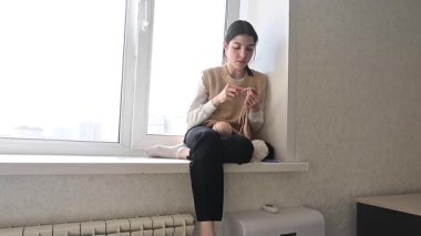 Genç beyaz kadın pencere eşiğinde otururken örgü örüyor.
