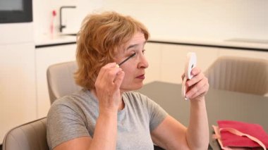 Yaşlı beyaz bir kadın kirpiklerine rimel sürüyor ve küçük bir aynaya bakıyor.