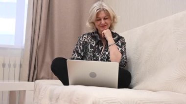 Kanepede otururken laptop kullanan olgun bir kadın.