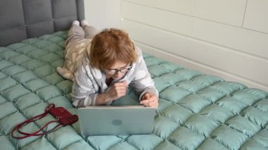 Yaşlı, beyaz, pijamalı bir kadın dizüstü bilgisayar kullanarak yatağa uzanıyor.