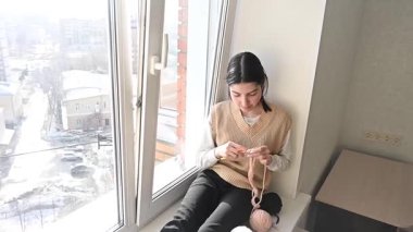 Genç beyaz kadın pencere eşiğinde otururken örgü örüyor.