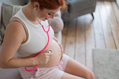 Hamile bir kadın fonendoskop kullanarak bebeğin nabzını dinler.
