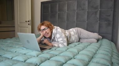 Pijamalı, yaşlı, beyaz bir kadın yatağında dizüstü bilgisayardan video izliyor.